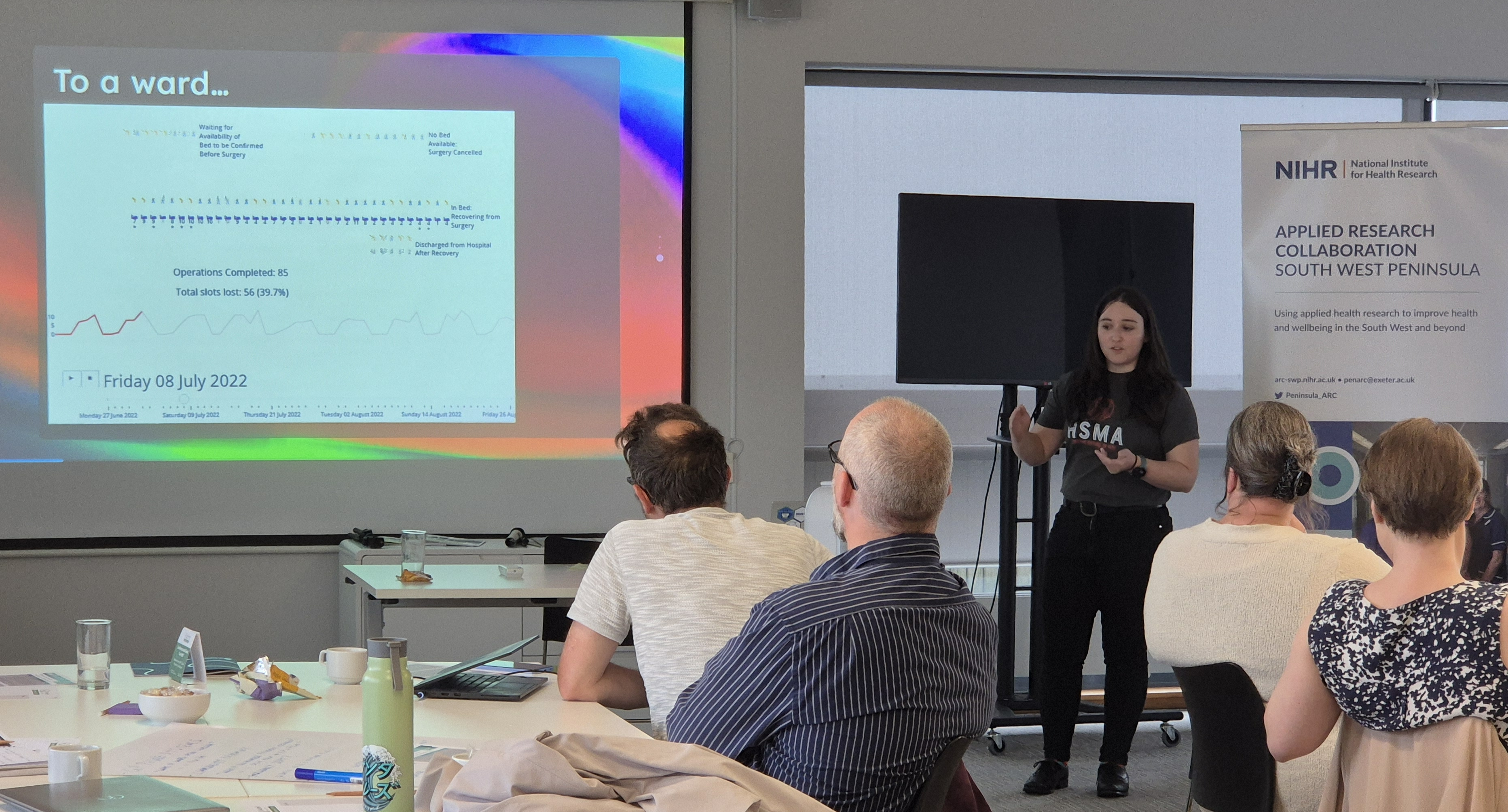 An image of the speaker in a HSMA t-shirt, talking to a room full of people about a simulation model of a hospital ward